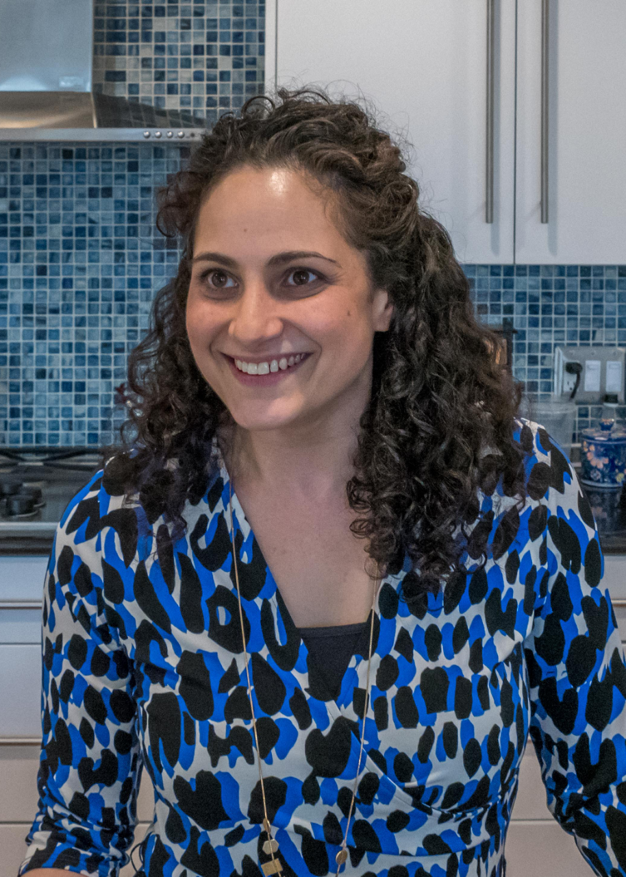 A smiling woman (Samantha Woll) with long brown curly hair, wearing a patterned blue, black and white dress, and standing in a white kitchen with dark and light blue mosaic tiled backsplash.