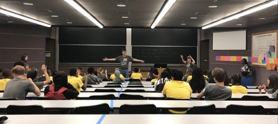In a photo taken from the back of the room, a male professor stands at the front of a U-M lecture room.  Students are seated at long tables facing the man and several other adults at the front of the room. The students are all listening intently, some raise their hands in the "jazz hands" gesture. The students, a mix of male and female, Black, white and POC, are wearing either yellow or gray t-shirts.