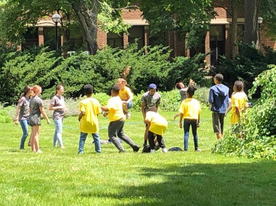 A group of laughing Math Corps campers (in yellow) and their TAs (in gray) stand in a circle on a U-M campus lawn.