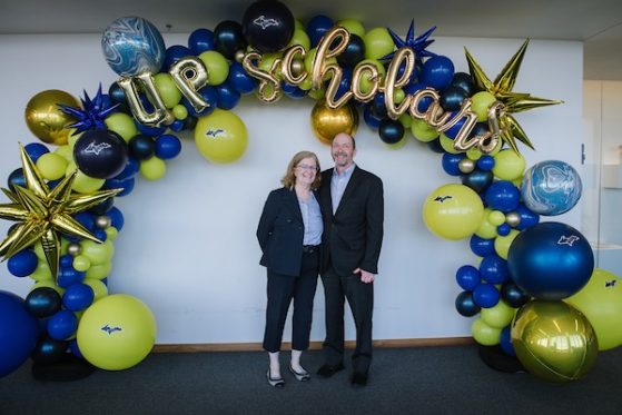 A couple (a man and a woman), each wearing a dark pantsuit with a light blue open-necked shirt. She is wearing glasses and has light reddish hair, his hair is close cropped and he is wearing glasses. They pose in front of a maize and blue balloon arch that reads U.P. Scholars.