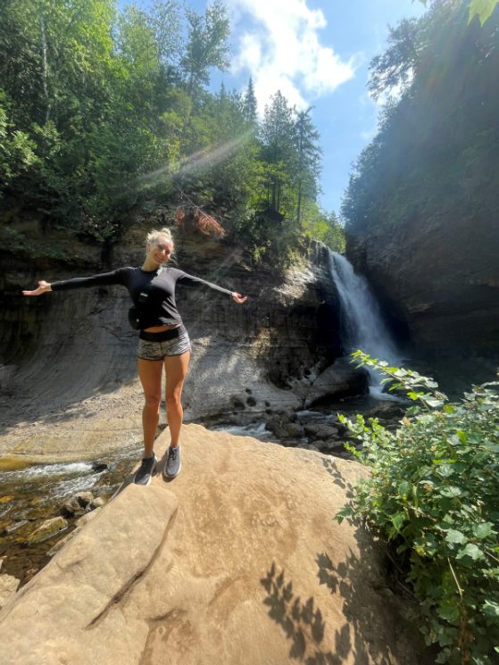 A student stands with her arms outstretched on a large boulder in front of pine trees and a waterfall. She is wearing running shorts, a long-sleeved top and hiking shoes. She has blond hair.