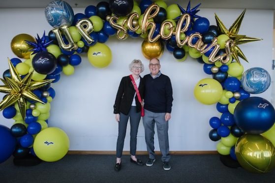 A couple (a man and a woman), each wearing gray pants a dark top, she has gray hair and is wearing glasses. She is carrying a red cross body bag with a red strap. He has thinning hair and is wearing glasses and blue shirt under his dark sweater. They pose in front of a maize and blue balloon arch that reads U.P. Scholars.