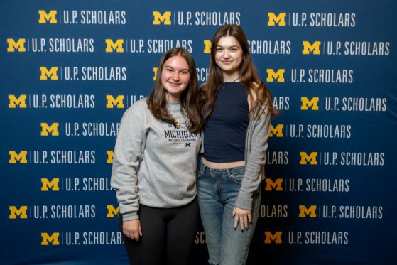 Two female students pose in front of a blue U.P. Scholars step and repeat background with the maize block M. They are both casually dressed, wearing jeans and sweatshirts, and smiling.
