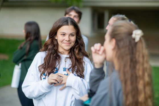 A student wearing a sweatshirt, with long dark wavy hair, stands smiling and fiddling with a ring on her middle finger. She is in conversation with another student in the foreground who has long light brown hair.