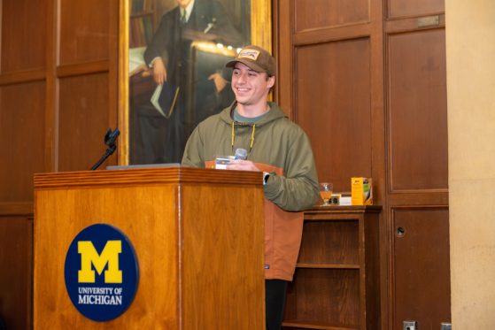 A male student stands at a wooden podium with a round blue sign that has the maize block M on the front. He is casually dressed in an olive and orange colored hooded sweatshirt, jeans and a brown baseball cap.