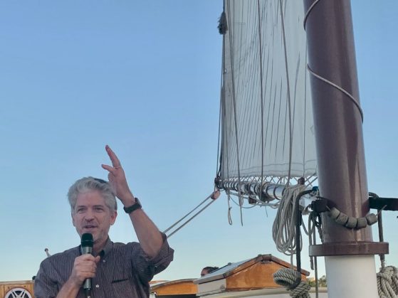 David Foster speaks into a microphone on the deck of a sailboat. He is wearing a button down shirt with sleeves rolled up to the elbow, dark khaki chino pants and a black wrist watch.