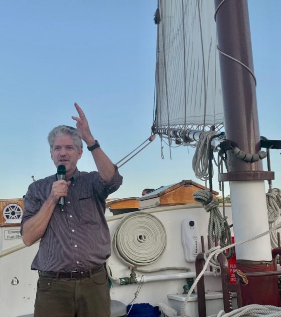 David Foster speaks into a microphone on the deck of a sailboat. He is wearing a button down shirt with sleeves rolled up to the elbow, dark khaki chino pants and a black wrist watch.