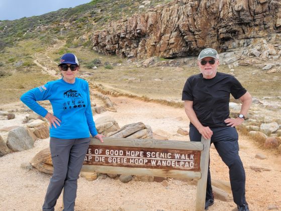 Sidonie (Sid) Smith and William (Bill) Kinley are pictured on a trip to South Africa. They pose on a hiking trail, each standing at either side of a sign that says "Cape of Good Hope Scenic Walk" in English and Afrikaans. Sid wears a cap, a blue long-sleeved shirt, gray pants, and hiking boots. Bill wears a black short-sleeved shirt, gray pants, and hiking boots.