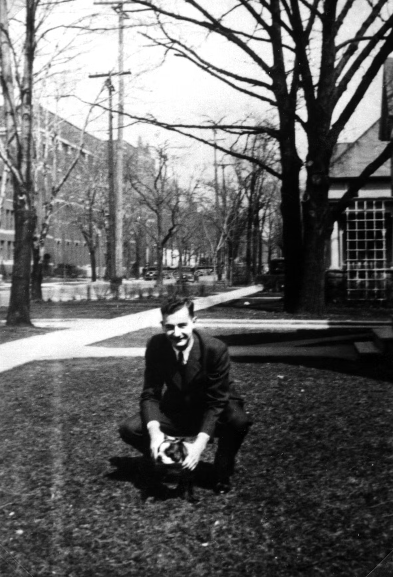 A historic (ca. 1930s) black and white image of a smiling young man crouching on a campus street with a small bulldog.