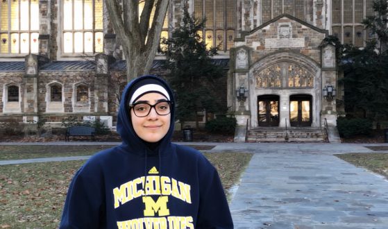 Noor is pictured in a navy blue Michigan Wolverines sweatshirt. She stands in the center of the Law Quad, fallen leaves cover the grass and the U-M Law Library is in the background.
