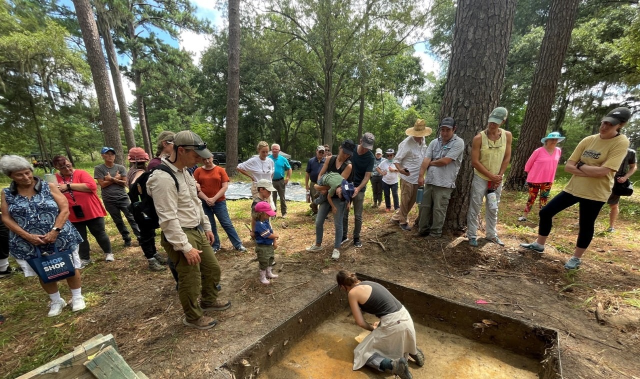 A crowd of people are gathered around a shallow pit at an archaeological dig site in South Carolina.