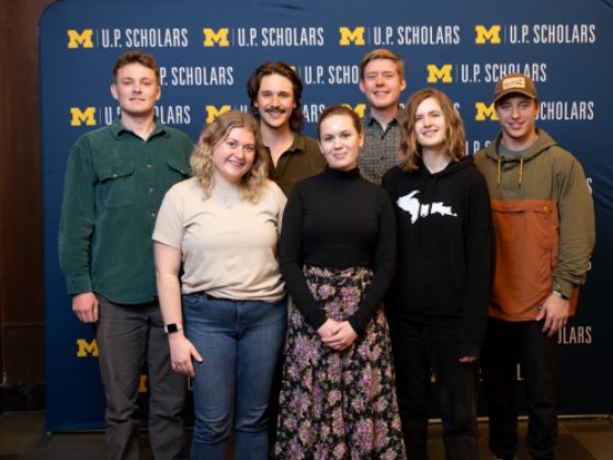 A group of seven students stands smiling in two rows. The three female students stand in the front row, the four male students stand in the back row. They stand in front of a blue U.P. Scholars step and repeat background with a maize block M. They are casually dressed in jeans and shirts with the one student in the front row wearing a black long-sleeved top and floral skirt. 