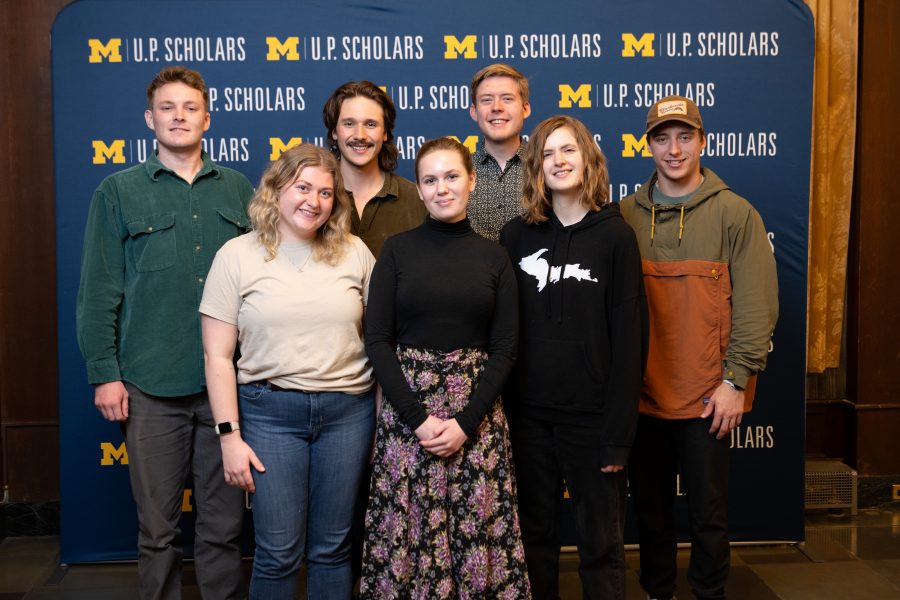 A group of seven students stands smiling in two rows. The three female students stand in the front row, the four male students stand in the back row. They stand in front of a blue U.P. Scholars step and repeat background with a maize block M. They are casually dressed in jeans and shirts with the one student in the front row wearing a black long-sleeved top and floral skirt. 