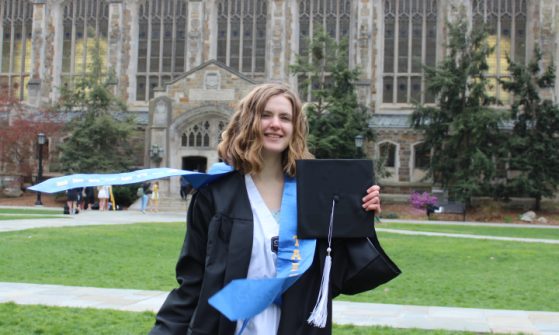 A female student with shoulder length wavy brown hair stands smiling in the Law Quad. She is wearing a black graduation gown with a light blue stole that is blowing in the wind. She holds a black graduation cap with a white tassel.