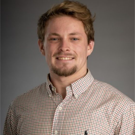 A male student with light brown hair and a brown goatee and mustache smiles in a professional headshot. He is wearing a green and red checkered Polo button down dress shirt.