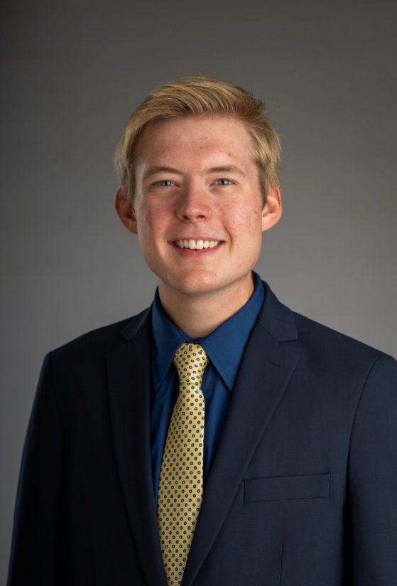 A male student smiles in a professional headshot. He has blond hair. He is wearing a dark blue dress shirt, yellow and blue tie, and a navy blue suit coat.