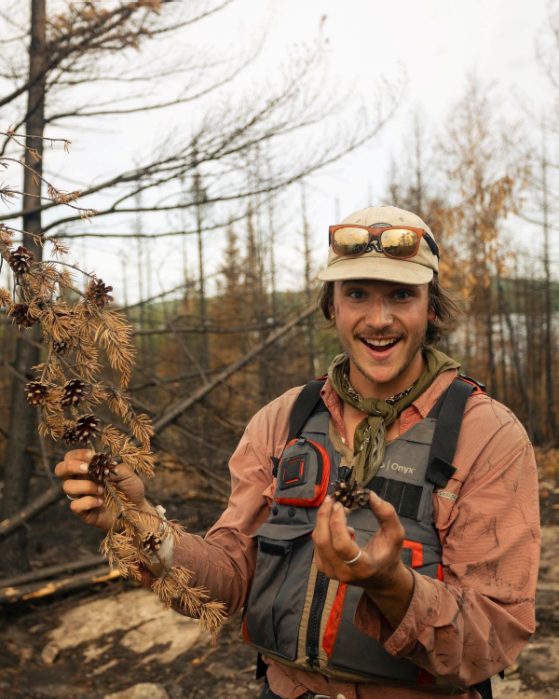 A student stands in the forest holding a pine cone, next to a pine tree. He is smiling and wearing hiking gear, including a vest with many pockets, a neck scarf, cap and has sunglasses on the top of his head. 