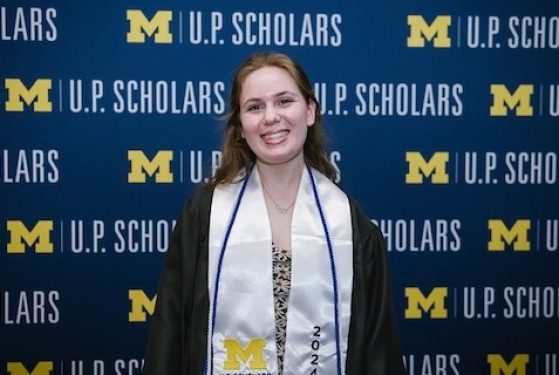 A female student with long wavy brown hair that is pinned back, stands in front a blue U.P. Scholars step and repeat background with the maize block M. She wears a black graduation gown with a white U.P. Scholars Class of 2022 stole and a navy blue chord.
