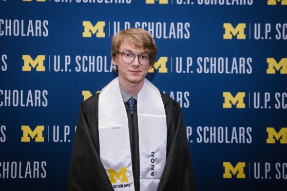 A male student with blond hair and glasses stands in front a blue U.P. Scholars step and repeat background with the maize block M. He wears a black graduation gown with a white U.P. Scholars Class of 2024 stole.