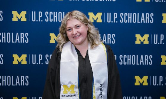 A female student with long wavy blond hair, stands in front a blue U.P. Scholars step and repeat background with the maize block M. She wears a black graduation gown with a white U.P. Scholars Class of 2024 stole.