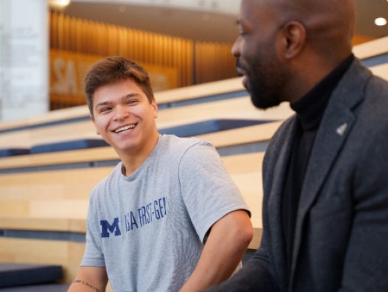 A student wearing a gray t-shirt with the words M First-Gen and an administrator in a dark suit sit in the LSA building having a conversation.