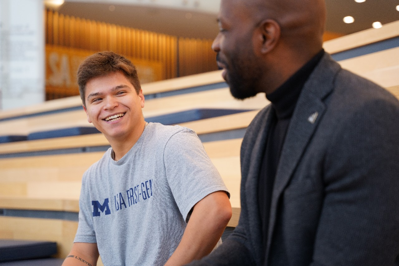 A student wearing a gray t-shirt with the words M First-Gen and an administrator in a dark suit sit in the LSA building having a conversation.