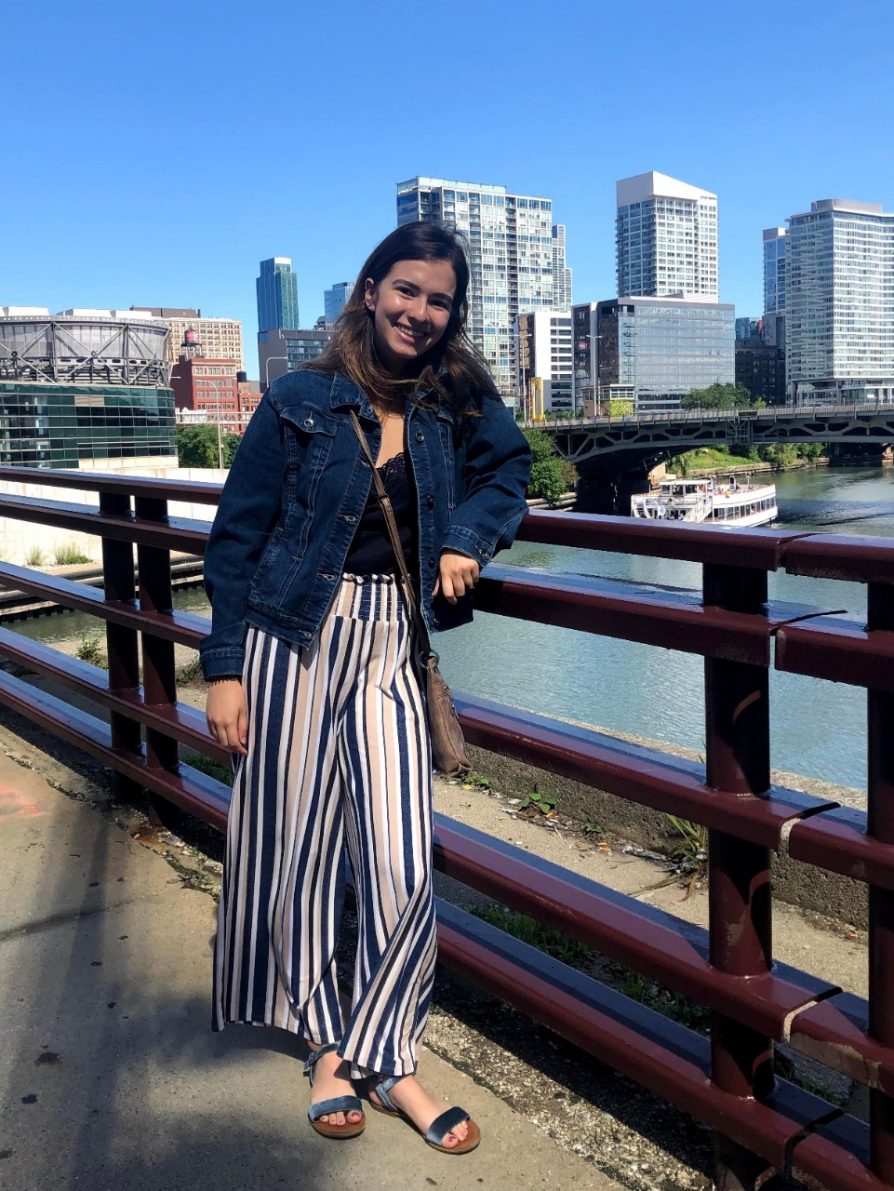 LSA student Athena Bryer is pictured smiling and facing the camera while standing on a bridge with a waterway, cityscape, and brilliant blue sky in the background.