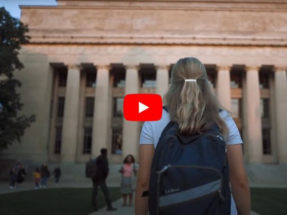 A student wearing a backpack is pictured walking toward, and looking up at, Angell Hall which is visible in the background. A “play” button is visible in the foreground indicating this is a video link.