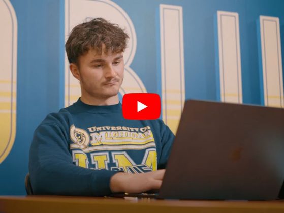 LSA student Alex Mamonov is pictured working on a laptop with a large scale “Go Blue” stenciled on a wall behind him. A “play” button can be seen in the foreground indicating that this is a link to a video.