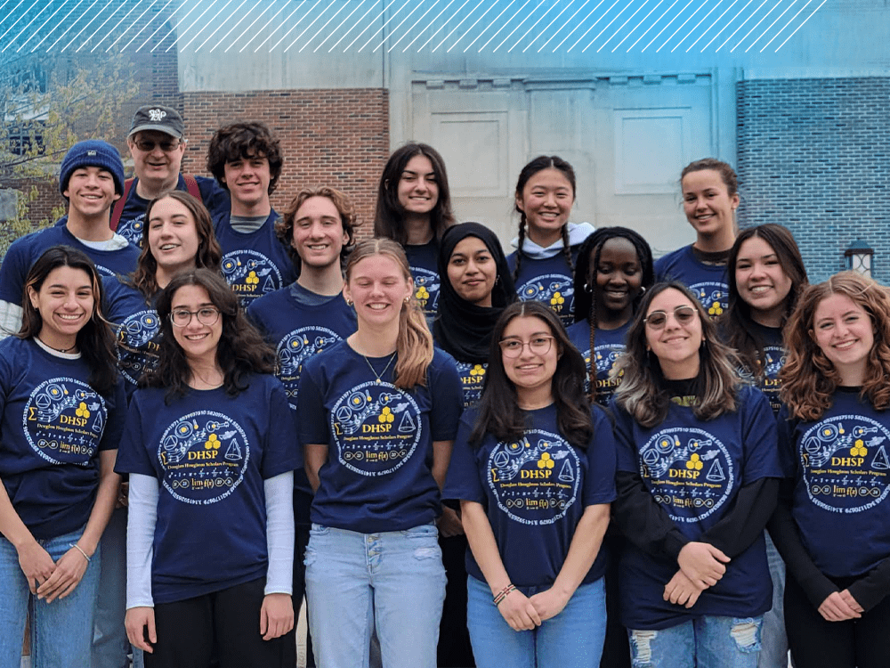 A group of 16 students, arranged in three rows, poses on the steps of a university building. Each person wears matching navy blue short-sleeved t-shirts with the letters DHSP in yellow, within a circular design that features equations and other recognizable math symbols. DHSP director Mark Conger is in the back row at the far left.