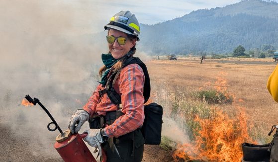 Rachel, in a hard hat, eye protection, a flannel shirt, and other firefighting gear, poses in a dry field with a small fire in the background.