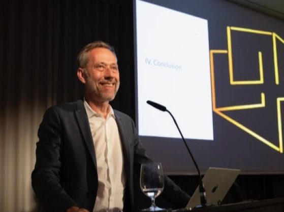 Man with grey hair and beard stands behind a podium with a projection screen to his right