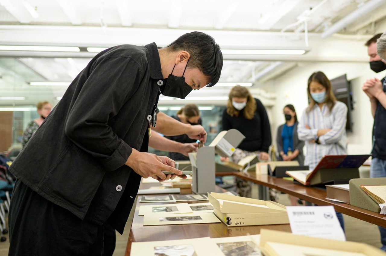 The artist, leaning over a table containing artifacts, examines a photograph from the museum's collection.