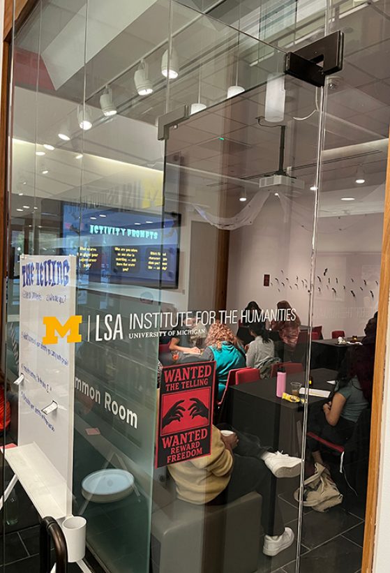 The image shows a glass-walled room at the LSA Institute for the Humanities, University of Michigan. Inside, people are seated at tables, engaged in an activity. A poster with “Wanted The Telling” and “Reward Freedom” is visible. 