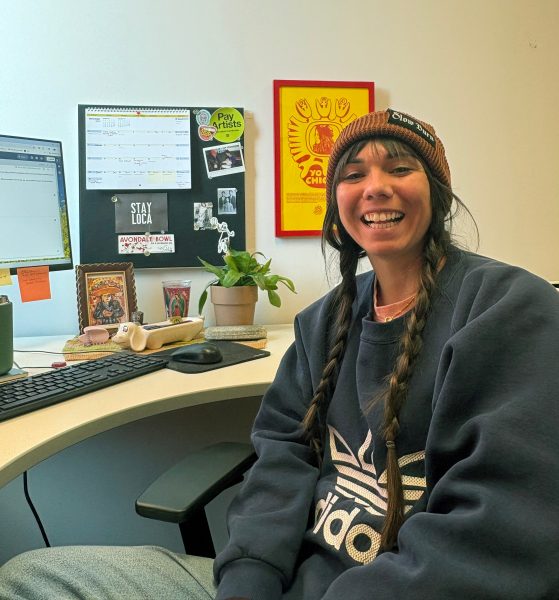 Julianna Loera-Wiggins with long braids and a beanie smiles at a desk with a computer, a bulletin board, and plant.