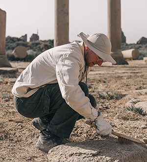 IPAMAA student Julius Arnold with brush in hand crouching to clear dirt from a stone 