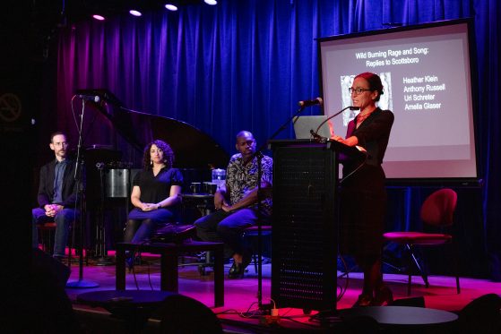 Four performers on stage: three sitting and one standing at a podium in front of a projection screen listing all four performer's names: Amelia Glaser, Heather Klein, Anthony Russell, and Uri Schreter