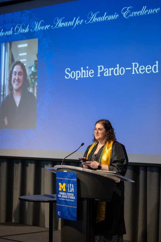 A graduate with dark curly hair stands smiling at a podium to accept the Deborah Dash Moore Award for Academic Excellence