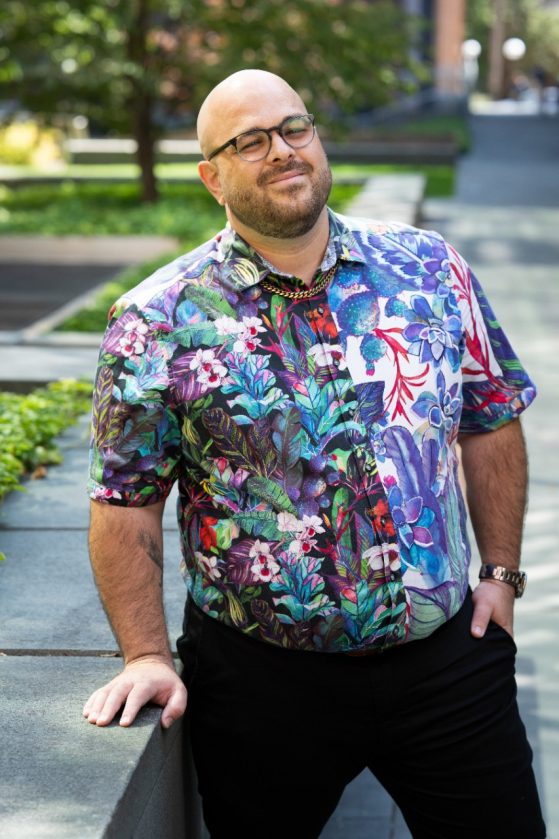 A bald man with glasses wearing a multicolored shirt and black pants leans against a wall smiling for a professional headshot