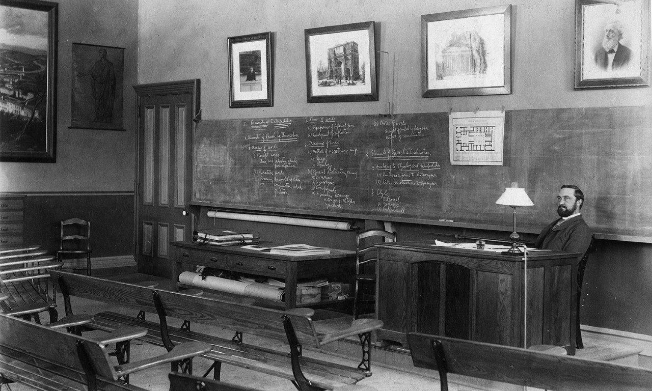 Nineteenth-century classroom showing Francis Kelsey sitting at a desk in front of a chalkboard covered in writing.