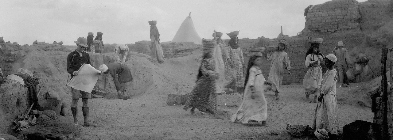 Scene of archaeological work in Egypt. At the left, researcher Enoch E. Peterson stands looking at a plan. Egyptian workers carry baskets on their heads in front of the mudbrick walls of Karanis.