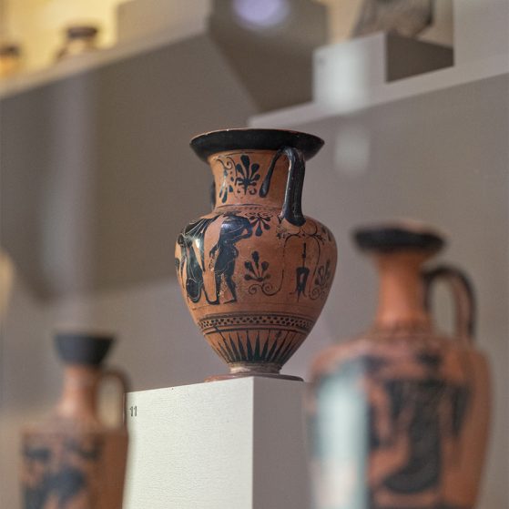 Glass display case at the Kelsey Museum containing several examples of Greek pottery. Prominently featured is a two-handled black-figure vase.