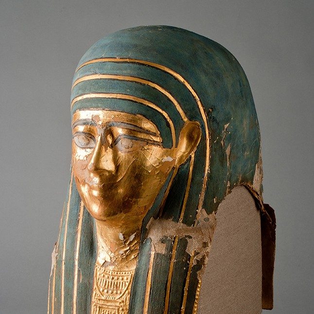 Vibrantly painted Egyptian mummy mask of a male face. The face and chest are gold, while the man’s long hairstyle is blue.