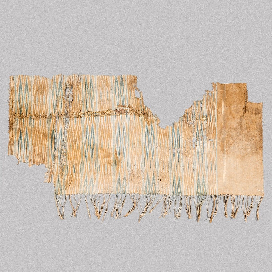 Blue and tan patterned textile with gold a line of text across the upper portion and fringe along the bottom.