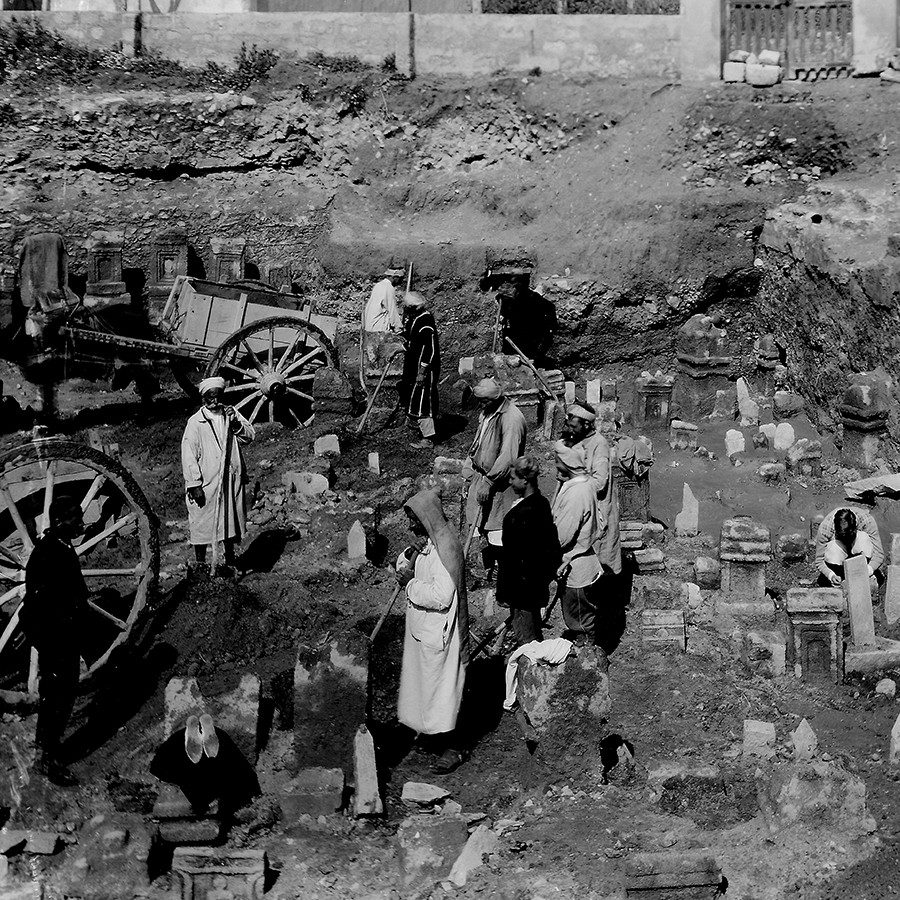 Archival photo showing a group of Tunisian men excavating a cemetery using shovels and carts. Several large stone stelae are visible, emerging from the dirt.