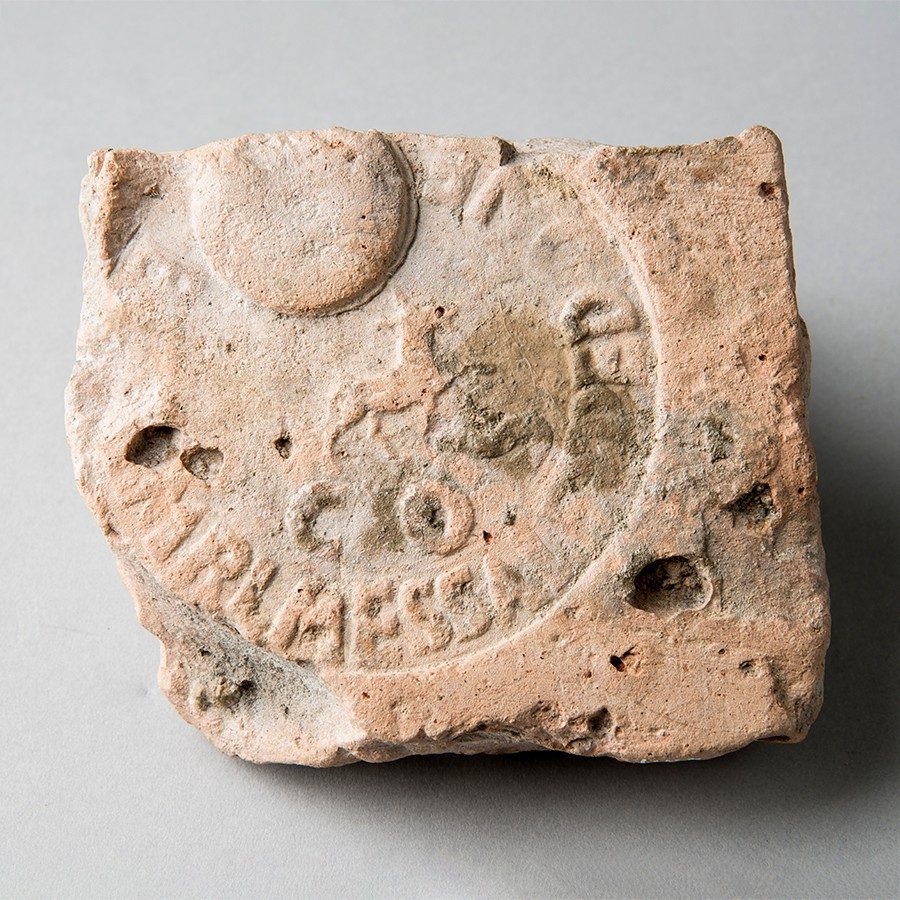 Lunette stamp on small square brick made of light reddish-brown clay. In the center, a wolf faces right., encircled by a two-line, circumferential inscription.