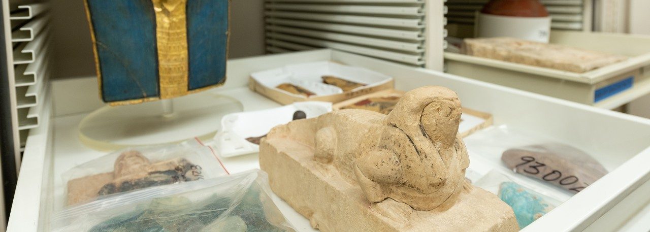 White Kelsey Museum storage cabinet. The door is open and a shelf pulled out, containing a gold and blue Egyptian mummy mask, a statue of Soknopaios, and other Egyptian objects.