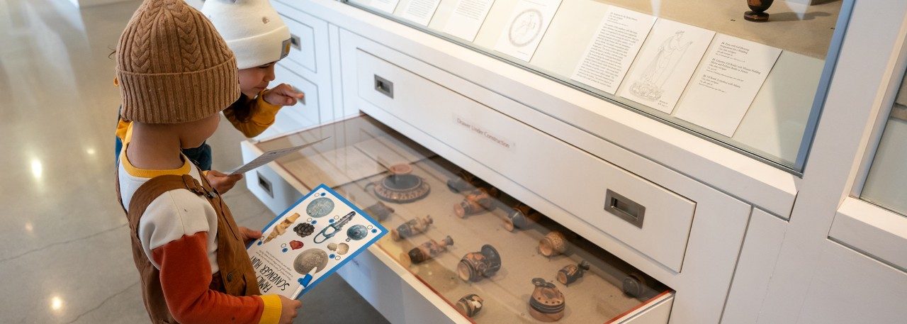 Two children look in a drawer containing Greek pottery to complete a scavenger hunt.