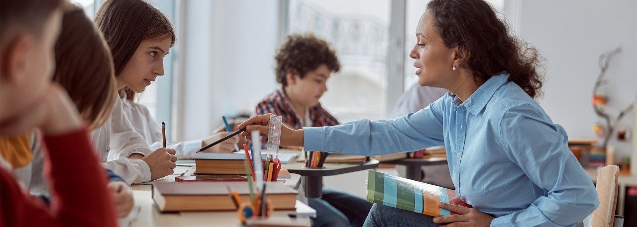 An elementary school teacher sits in a chair in front of an arc of student desks. She holds a book and points a colored pencil at a student’s work.