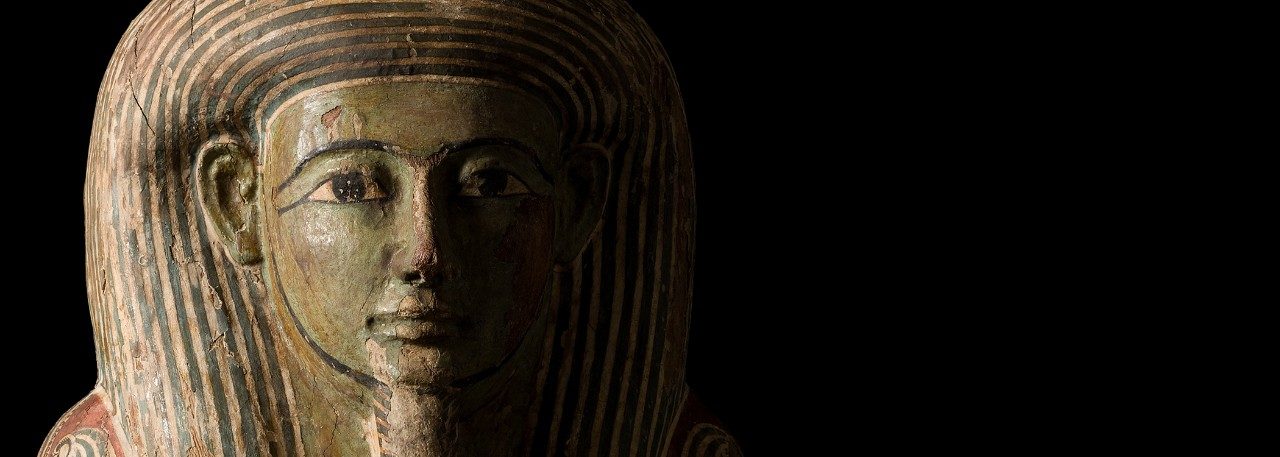 Close-up of the coffin lid of the Egyptian priest Djehutymose, a wooden artifact with a painted green face, curved eyebrows, eyes lined with kohl, a goatee, and blue- and white-striped headdress. 